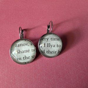 May include: A pair of round, silver-toned earrings. Each earring features a clear, domed glass over a circular image of text from a book. The text is in black on a white background. The earrings are displayed on a pink surface.