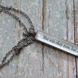 Puede incluir: Collar plateado con un colgante rectangular. El colgante presenta el texto "She was okay now, but h" en negro, con fuente serif. La cadena es de estilo eslabón clásico.