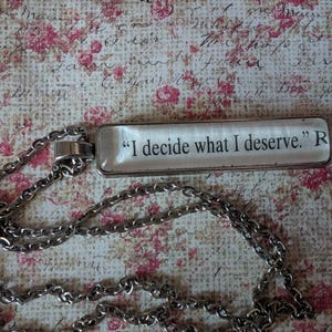 May include: A silver-toned necklace with a rectangular pendant. The pendant has the quote "I decide what I deserve." in black text. The chain is a linked design. The background is a floral pattern with pink and red roses.