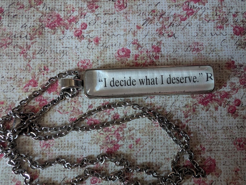 May include: A silver-toned necklace with a rectangular pendant. The pendant has the quote "I decide what I deserve." in black text. The chain is a linked design. The background is a floral pattern with pink and red roses.