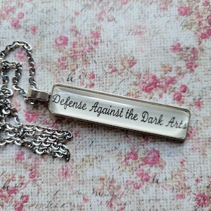 May include: A silver chain necklace with a rectangular pendant that says "Defence Against the Dark Arts" in black lettering.