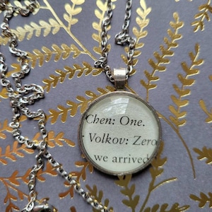 May include: A silver chain necklace with a round glass pendant. The pendant has the text "Chen: One. Volkov: Zero. We arrived." printed on it.