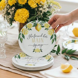May include: White paper plates with a lemon design and the text "She found her MAIN squeeze." The plates are decorated with lemons and green leaves. A hand is holding one plate, with a stack of plates below. Yellow flowers and lemons are in the background.