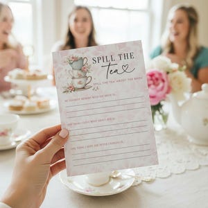 Op de afbeelding: Een roze en witte vrijgezellenfeest-spelkaart met de tekst "Spill the Tea" en een illustratie van een stapel theekopjes. De kaart heeft lijnen om op te schrijven, met een bloemenontwerp en een theekopje en theepot op de achtergrond.
