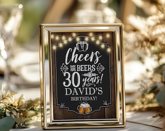 May include: A gold-framed sign with a rustic wood background and a chalkboard overlay. The sign reads "Cheers and Beers to 30 years! David's Birthday!" with string lights and beer mugs.