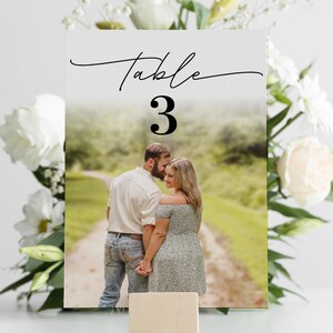 Puede incluir: Una tarjeta con el n&uacute;mero de mesa de boda con la palabra "Table" en escritura elegante y el n&uacute;mero "3" en negrita. La tarjeta presenta una foto de una pareja y est&aacute; sostenida por un peque&ntilde;o bloque de madera, rodeado de flores blancas.