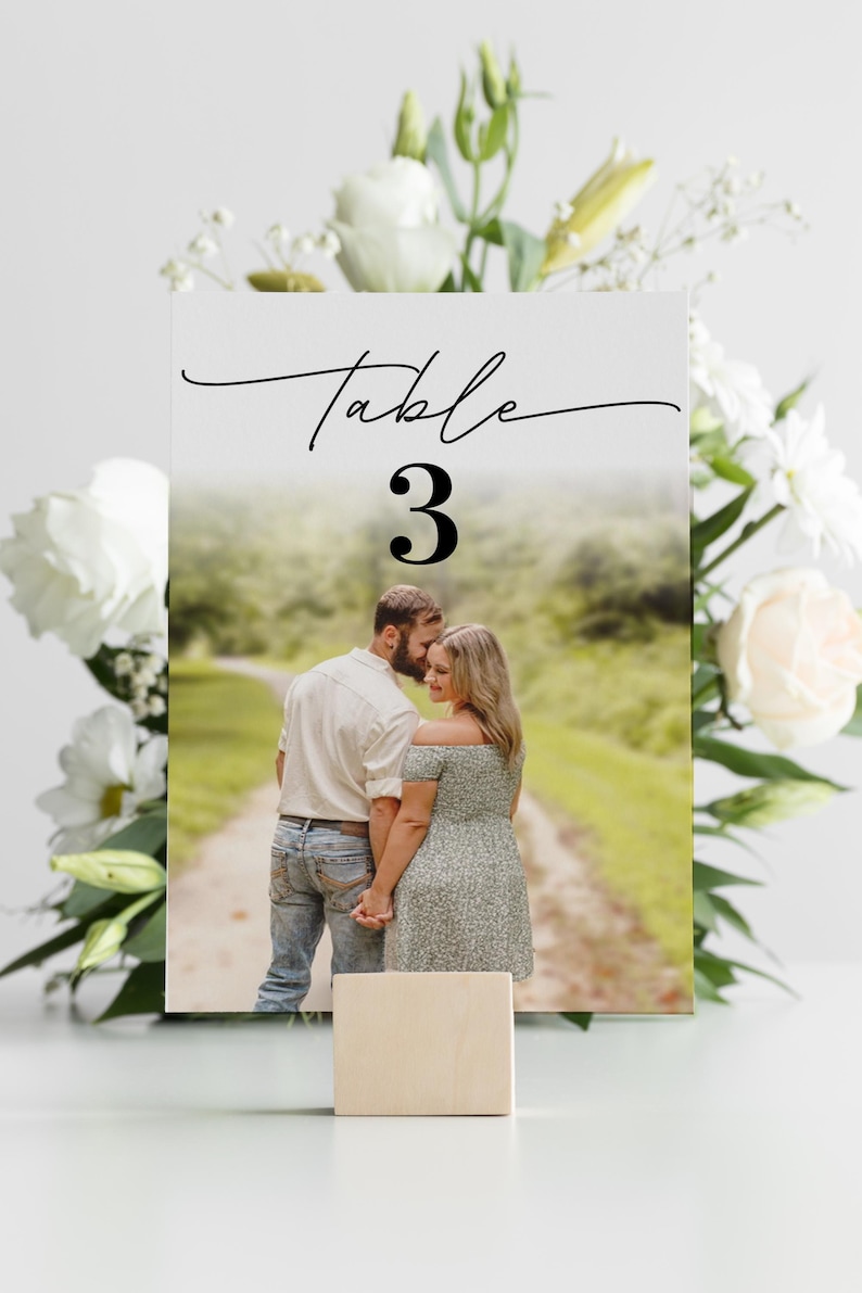 Puede incluir: Una tarjeta con el n&uacute;mero de mesa de boda con la palabra "Table" en escritura elegante y el n&uacute;mero "3" en negrita. La tarjeta presenta una foto de una pareja y est&aacute; sostenida por un peque&ntilde;o bloque de madera, rodeado de flores blancas.
