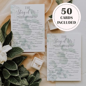K&ouml;nnte beinhalten: Zwei wei&szlig;e Karten mit dem Text "The Story of Us" und Ausf&uuml;llaufforderungen, mit floralem Design. Ein runder Aufkleber lautet "50 KARTEN ENTHALTEN". Eine Ringbox mit einem Ring ist ebenfalls sichtbar.