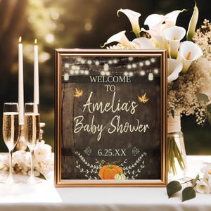 May include: A rustic brown welcome sign with a gold frame, featuring the text "Welcome to Amelia's Baby Shower" with a string of lights and fall leaves. The sign also includes a pumpkin and a small white gourd.