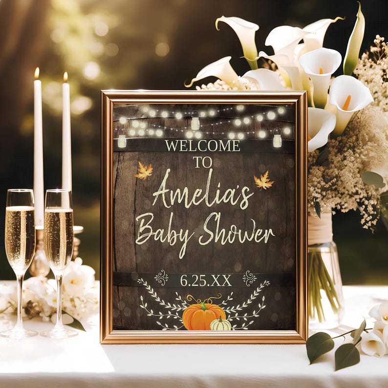 May include: A rustic brown welcome sign with a gold frame, featuring the text "Welcome to Amelia's Baby Shower" with a string of lights and fall leaves. The sign also includes a pumpkin and a small white gourd.