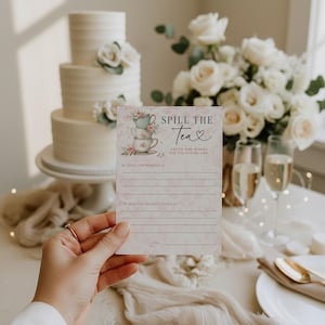 Op de afbeelding: Een hand houdt een roze en witte "Spill the Tea" advieskaart voor een bruidsfeest. De kaart toont gestapelde theekopjes en bloemmotieven. Een drielaagse taart en een boeket witte rozen staan op de achtergrond.