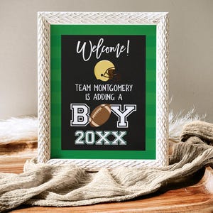 May include: A white framed sign with a green background and black text. The sign says "Welcome! Team Montgomery is adding a boy 20XX". There is a football and a football helmet on the sign.