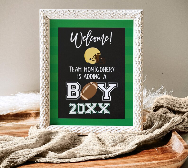 May include: A white framed sign with a green background and black text. The sign says "Welcome! Team Montgomery is adding a boy 20XX". There is a football and a football helmet on the sign.