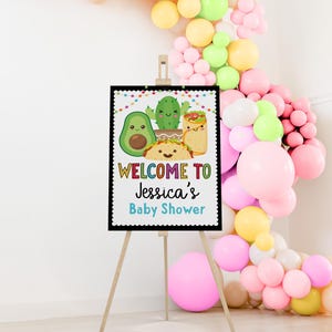 May include: Welcome to Jessica's Baby Shower sign with a black background and colorful text. The sign features a cartoon avocado, cactus, and taco with a burrito.