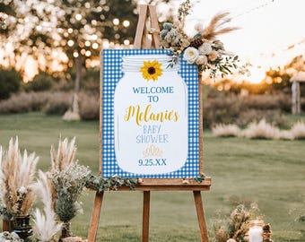 Op de afbeelding: Welkom bij Melanie's Baby Shower bord met een blauw-witte ruitjesachtergrond, een glazen pot met een zonnebloem en de datum 9.25.XX.