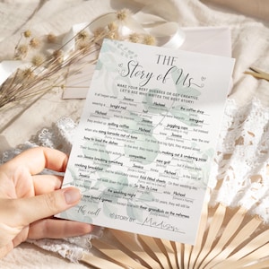 K&ouml;nnte beinhalten: Eine wei&szlig;e Hochzeitsspielkarte mit dem Titel "The Story of Us" und L&uuml;cken zum Ausf&uuml;llen. Die Karte hat ein Blumendesign und wird von einer Hand gehalten. Die Texte enthalten Aufforderungen wie "wearing a bright red" und "the wedding night".