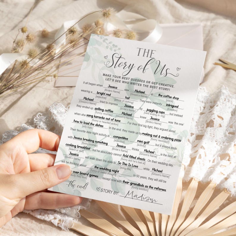 K&ouml;nnte beinhalten: Eine wei&szlig;e Hochzeitsspielkarte mit dem Titel "The Story of Us" und L&uuml;cken zum Ausf&uuml;llen. Die Karte hat ein Blumendesign und wird von einer Hand gehalten. Die Texte enthalten Aufforderungen wie "wearing a bright red" und "the wedding night".