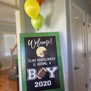 May include: A green and black welcome sign with the text "Welcome! Team Montgomery is adding a boy 2020". The sign has a football and a football helmet graphic. Three balloons are in the background, one yellow, one green, and one brown.
