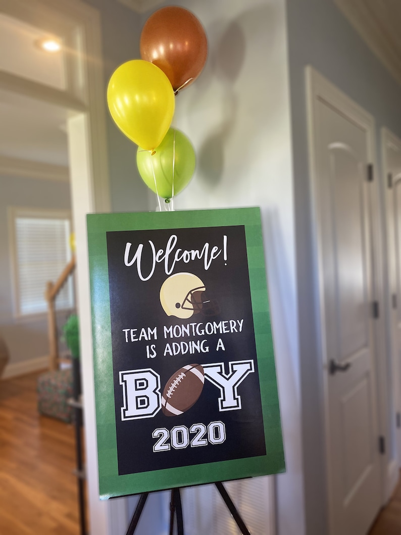 May include: A green and black welcome sign with the text "Welcome! Team Montgomery is adding a boy 2020". The sign has a football and a football helmet graphic. Three balloons are in the background, one yellow, one green, and one brown.