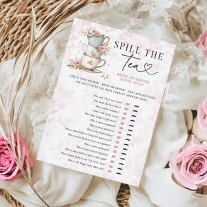 Op de afbeelding: Een roze en witte bruidsdouche-spelkaart met de tekst "Spill the Tea". De kaart heeft een stapel theekopjes en bloemaccenten. Roze rozen en gedroogde planten omringen de kaart.