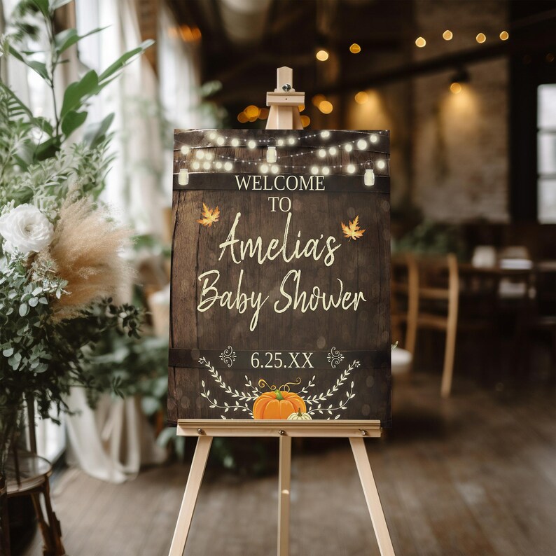 May include: A rustic wooden sign with a welcome to Amelia's baby shower message. The sign is decorated with fall leaves, pumpkins, and string lights. The date of the shower is 6.25.XX.