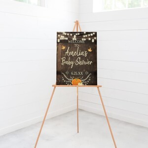 May include: Welcome to Amelia's Baby Shower sign with a rustic wood background, string lights, fall leaves, and pumpkins. The date is 6.25.XX.