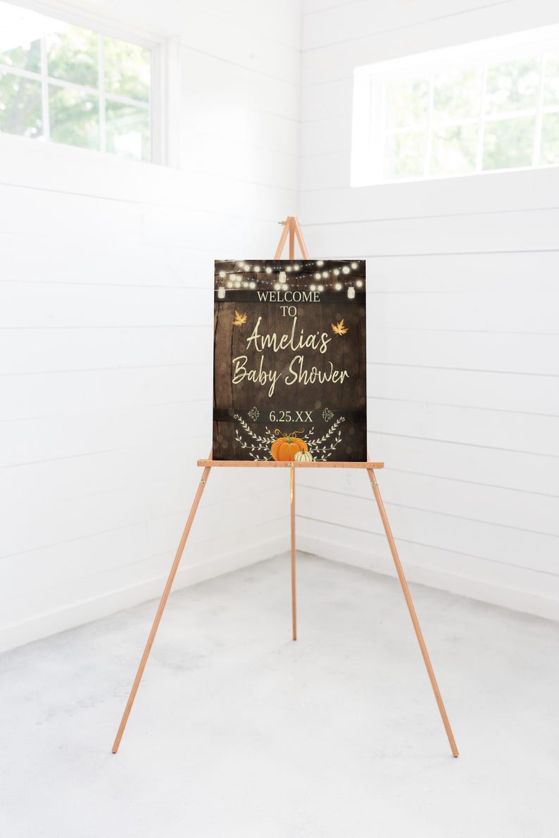 May include: Welcome to Amelia's Baby Shower sign with a rustic wood background, string lights, fall leaves, and pumpkins. The date is 6.25.XX.