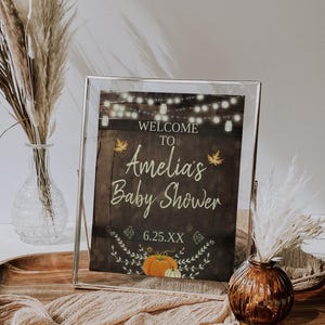 May include: A framed sign with a rustic wood background and a welcome to Amelia's Baby Shower message. The sign features a string of lights, fall leaves, and a pumpkin.