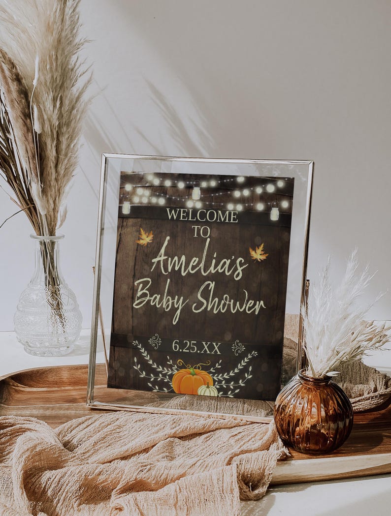 May include: A framed sign with a rustic wood background and a welcome to Amelia's Baby Shower message. The sign features a string of lights, fall leaves, and a pumpkin.