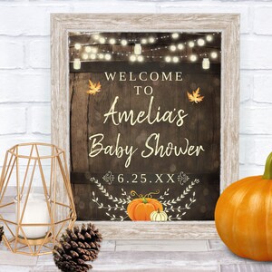 May include: A rustic wooden sign with a welcome to Amelia's Baby Shower message, featuring string lights, fall leaves, and pumpkins. The date 6.25.XX is also included.
