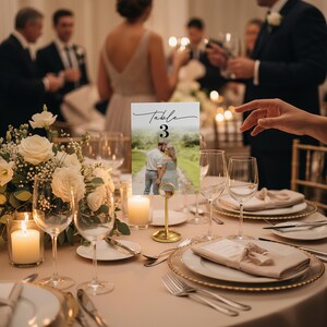 Puede incluir: Una mesa de recepci&oacute;n de bodas con un centro de mesa floral, velas y cubiertos. Un portatarjetas dorado muestra una tarjeta con el texto "Table 3" y una foto de una pareja. La mesa est&aacute; puesta con cristaler&iacute;a y servilletas.