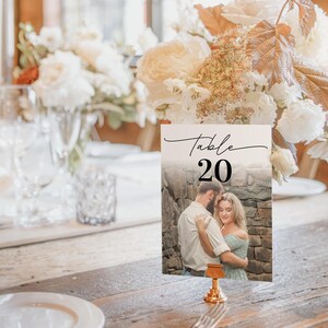 Puede incluir: Una tarjeta con el n&uacute;mero de mesa con el texto "Table 20" en escritura negra. La tarjeta presenta una foto de una pareja abraz&aacute;ndose. La tarjeta est&aacute; sujeta por un soporte de metal dorado y est&aacute; sobre una mesa de madera.