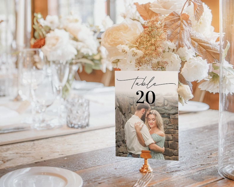Puede incluir: Una tarjeta con el n&uacute;mero de mesa con el texto "Table 20" en escritura negra. La tarjeta presenta una foto de una pareja abraz&aacute;ndose. La tarjeta est&aacute; sujeta por un soporte de metal dorado y est&aacute; sobre una mesa de madera.