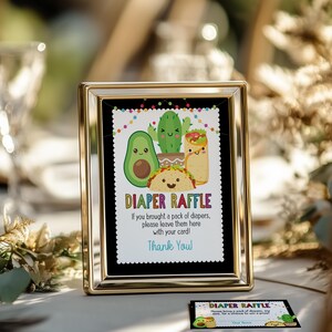May include: A gold-framed sign with a black background and colorful text that reads "Diaper Raffle". The sign features a cartoon avocado, cactus, and taco with smiling faces. The text below the image reads "If you brought a pack of diapers, please leave them here with your card! Thank You."