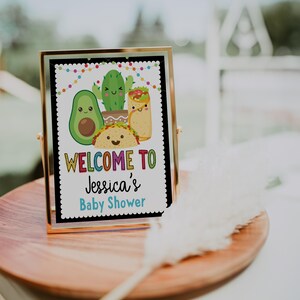 May include: A gold-framed sign with a black background and colorful text that reads "Welcome to Jessica's Baby Shower". The sign features a cartoon avocado, cactus, and taco.