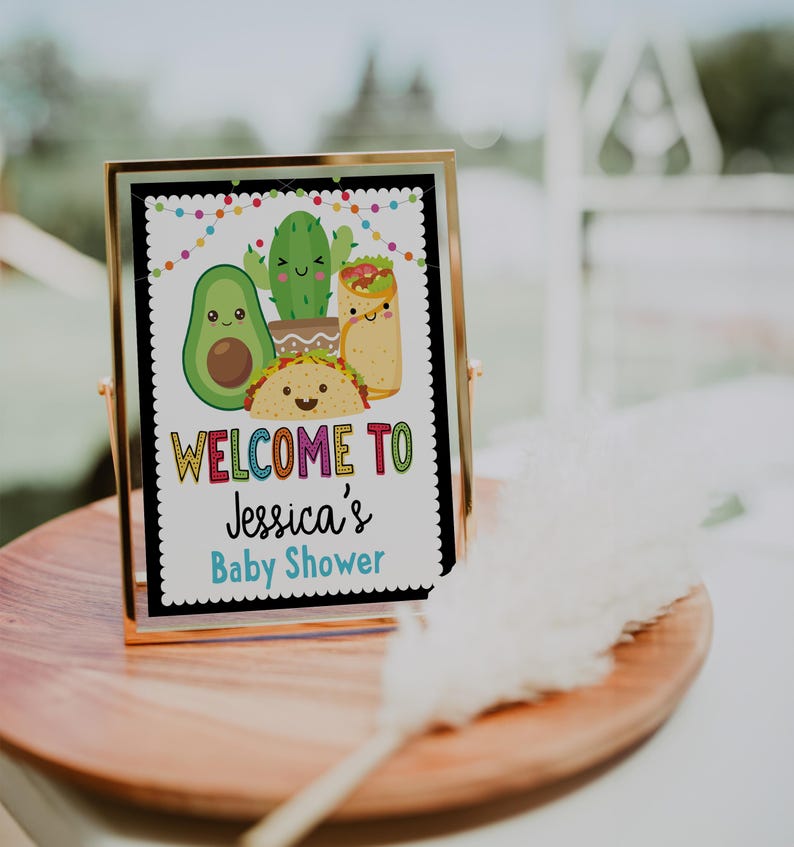 May include: A gold-framed sign with a black background and colorful text that reads "Welcome to Jessica's Baby Shower". The sign features a cartoon avocado, cactus, and taco.