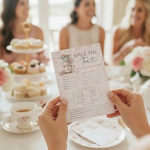Op de afbeelding: Een persoon houdt een roze en witte "Spill the Tea" spelkaart met bloemige theekopjes illustraties. De kaart is voor een theeparty, met invulvakken voor gasten. Andere theeparty-items zijn zichtbaar.