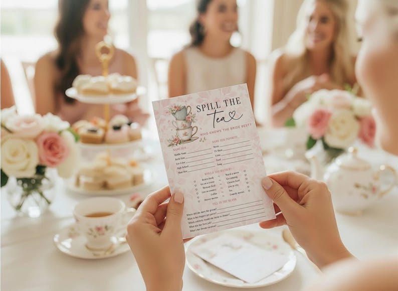 Op de afbeelding: Een persoon houdt een roze en witte "Spill the Tea" spelkaart met bloemige theekopjes illustraties. De kaart is voor een theeparty, met invulvakken voor gasten. Andere theeparty-items zijn zichtbaar.
