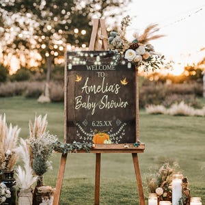 May include: A rustic wooden sign with a chalkboard surface, decorated with fall leaves and pumpkins. The sign reads "Welcome to Amelia's Baby Shower 6.25.XX".