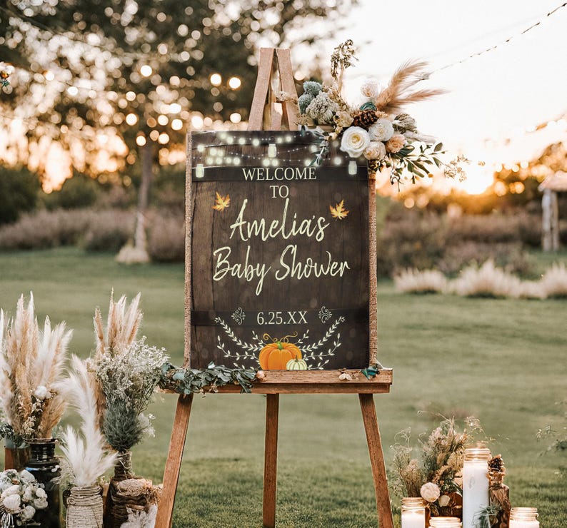 May include: A rustic wooden sign with a chalkboard surface, decorated with fall leaves and pumpkins. The sign reads "Welcome to Amelia's Baby Shower 6.25.XX".