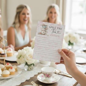 Op de afbeelding: Een bloemrijke bruidsdouche-spelkaart met de tekst "Spill the Tea" die voor een groep vrouwen wordt gehouden. De kaart bevat een stapel theekopjes en ruimte om advies te schrijven. Een dessertstandaard met meerdere niveaus bevat cupcakes.