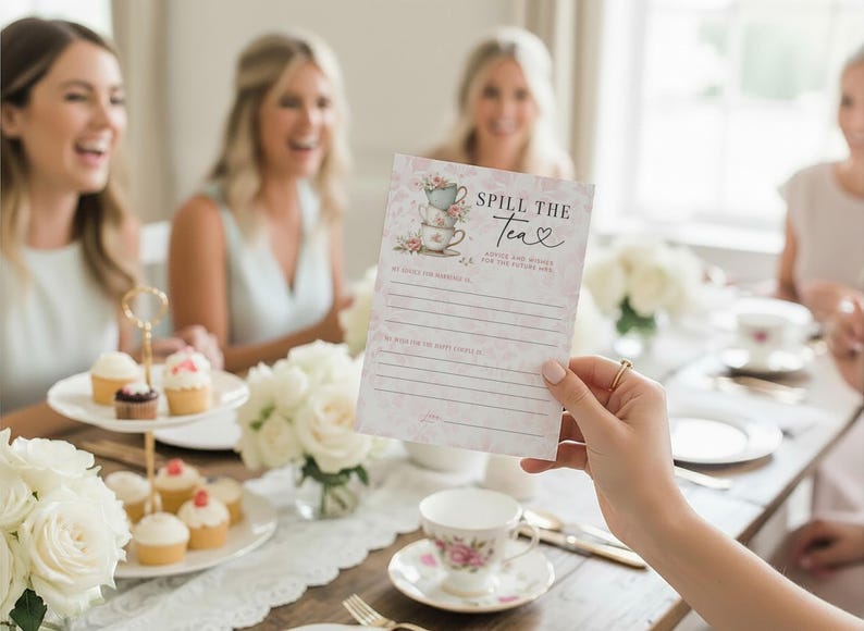 Op de afbeelding: Een bloemrijke bruidsdouche-spelkaart met de tekst "Spill the Tea" die voor een groep vrouwen wordt gehouden. De kaart bevat een stapel theekopjes en ruimte om advies te schrijven. Een dessertstandaard met meerdere niveaus bevat cupcakes.