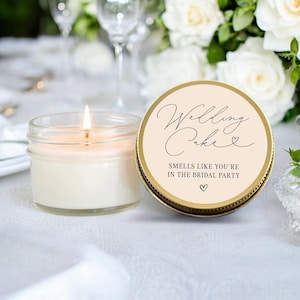 May include: A lit, white candle in a clear glass jar next to a candle lid. The lid is gold with the words "Wedding Cake" and "Smells like you're in the bridal party" in black script. White flowers and glasses are in the background.