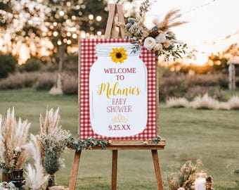 Op de afbeelding: Welkom bij Melanie's Baby Shower bord met een rood-wit geruite achtergrond en een glazen pot met een zonnebloem erop. De datum 9.25.XX staat op het bord gedrukt.
