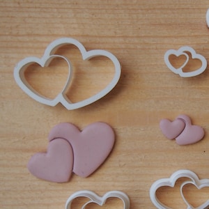 Pode incluir: Um conjunto de nove cortadores de biscoitos de plástico branco em forma de dois corações entrelaçados. Os cortadores são de vários tamanhos e estão dispostos sobre uma superfície de madeira marrom claro.