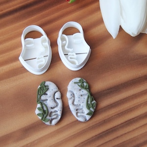 May include: Two white plastic cookie cutters shaped like faces with leaves around them, and two white plastic cookies with green leaves around them.