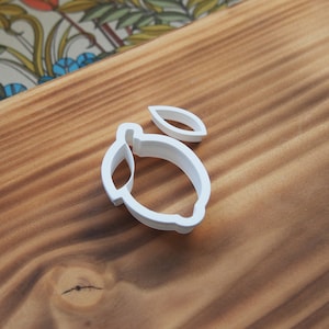 Könnte beinhalten: Ein weißer Ausstecher in Zitronenform aus Kunststoff mit einem separaten Blattteil. Der Ausstecher befindet sich auf einer Holzoberfläche mit Holzmaserung. Der Hintergrund zeigt eine farbenfrohe Blumentapete.