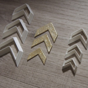 May include: Five sets of layered chevron-shaped cookie cutters in different sizes. Three sets are white and two sets are gold glitter.