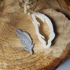 Könnte beinhalten: Ein weißer Plastik-Plätzchenausstecher in Form einer Feder auf einer Holzoberfläche. Eine graue Feder liegt ebenfalls auf der Oberfläche.