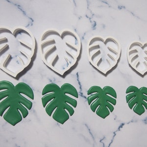 Puede incluir: Conjunto de cuatro cortadores de galletas de plástico blanco con forma de hojas de monstera, y seis hojas de monstera de fondant verde.